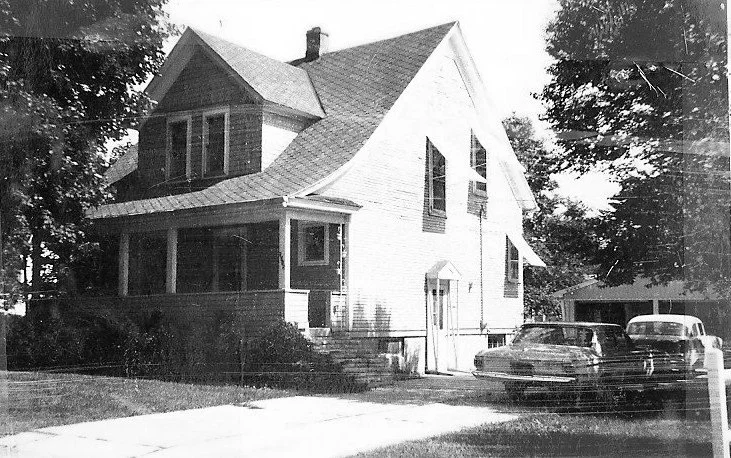 Black-and-white photo of a two-story house with a covered front porch, driveway and two vintage cars parked beside it.