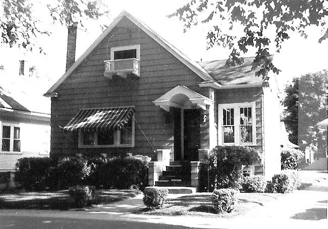 Black-and-white photo of a small gabled bungalow with front steps, striped window awning, upper balcony box, and shrubs lining the yard.