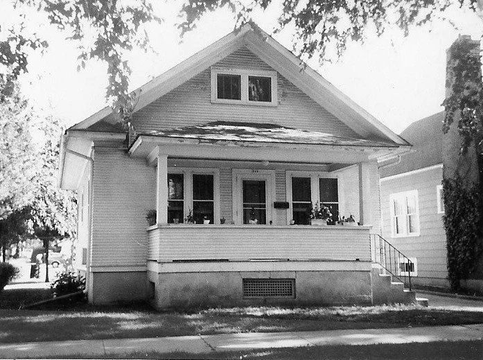 Black-and-white photo of a small bungalow with a covered front porch, three front windows, concrete steps at right, and trees framing the scene.