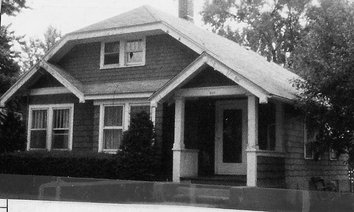 Black-and-white photo of a single-story Craftsman bungalow with a covered front porch, tapered columns, gabled roof, and shrubs in front.