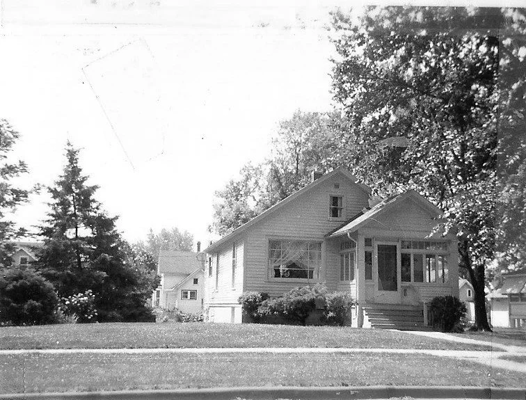 Black-and-white photo of a single-story house with a front porch, lawn, sidewalk, and large trees.