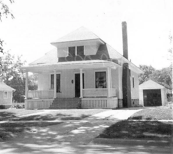 Black-and-white photo of a two-story bungalow with a wide front porch, tall chimney, driveway, and a detached garage to the right.