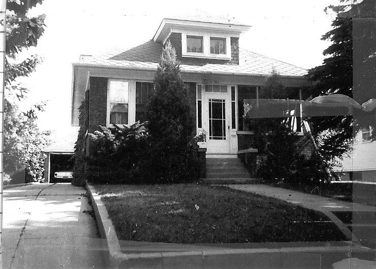 Black-and-white photo of a brick bungalow with front steps, shrubs, a small lawn and a driveway leading to a garage.