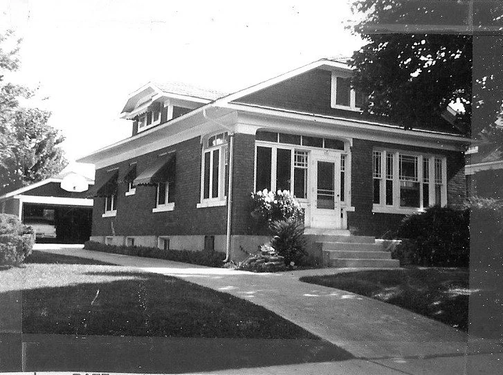 Black-and-white photo of a one-story brick bungalow with enclosed front porch, concrete walkway, lawn, and detached garage.