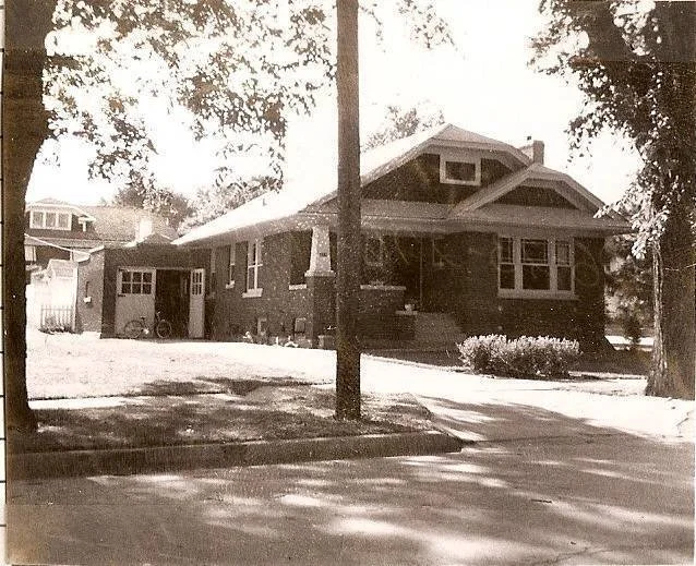 Sepia-toned photo of a single-story brick bungalow with a front porch, detached garage, and shaded tree-lined street.