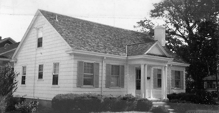 Black-and-white photo of a single-story clapboard house with a gabled roof, front porch with columns, and shuttered windows.