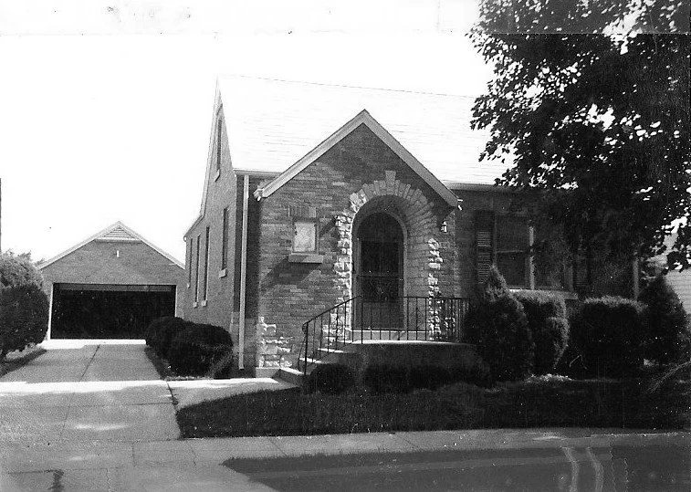 Black-and-white photo of a one-story brick house with an arched front doorway, small porch with railings, shrubs, and a detached garage.
