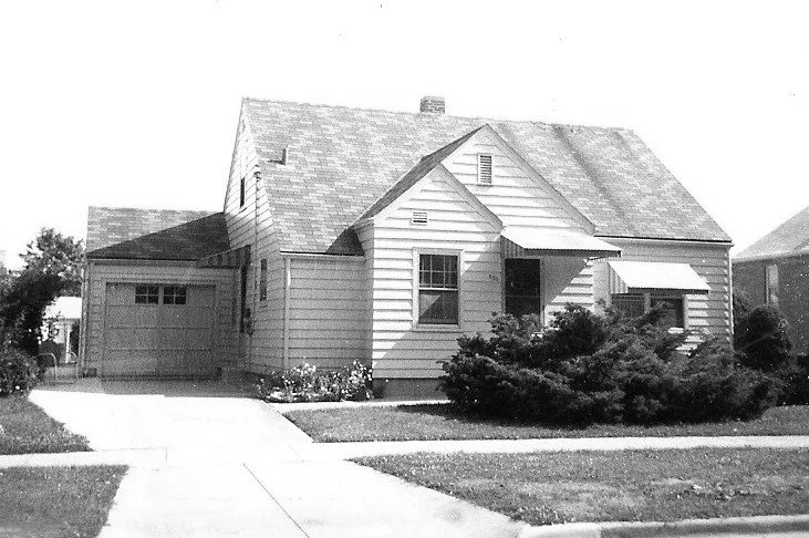 Black-and-white photo of a single-story wood-sided suburban house with attached garage, driveway, small front porch and front shrubs.