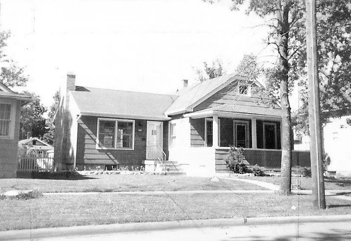 Black-and-white photo of a single-story bungalow with front porch, lawn, sidewalk, and a tree by a utility pole.