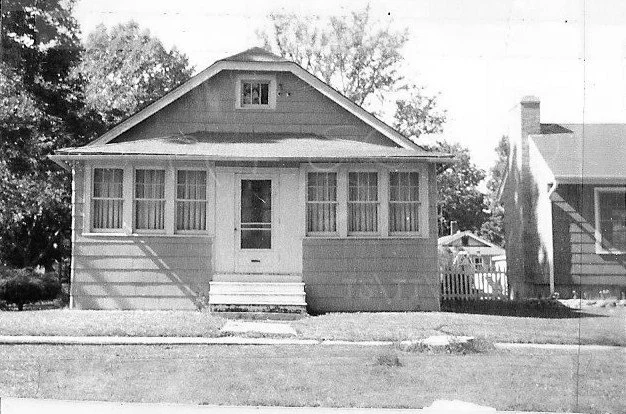 Black-and-white photo of a small single-story bungalow with an enclosed front porch, centered door and windows, and a front lawn.