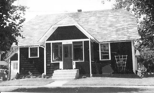 Black-and-white photo of a small one-story house with a covered front porch, three front windows, steps up from a lawn and trees at the edges.