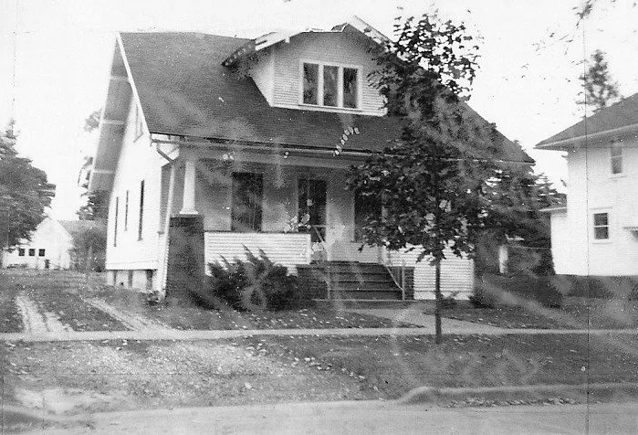 Black-and-white photo of a vintage two-story bungalow with a front porch, small tree and steps leading from the sidewalk.