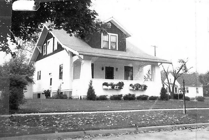 Black-and-white photo of a two-story Craftsman-style house with a wide front porch, window boxes, lawn, and sidewalk.