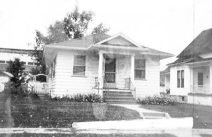 Black-and-white photo of a small single-story bungalow with a front porch supported by columns, steps, and a curved walkway through a grassy yard.