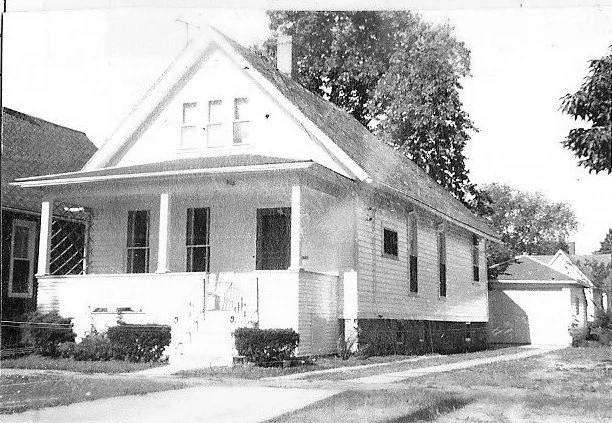 Black-and-white photo of a small gabled bungalow with a covered front porch, steps, shrubs, and a driveway leading to a detached garage.