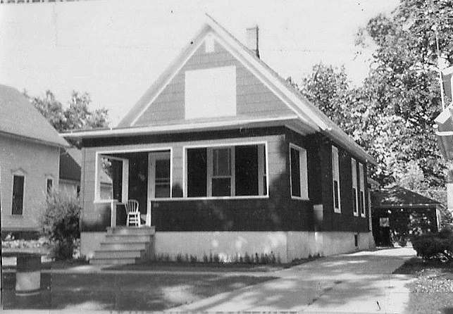 Black-and-white photo of a single-story bungalow with a screened front porch, a chair on the steps, driveway to the right, and trees in the background.