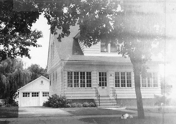 Black-and-white photo of a two-story house with an enclosed front porch, detached garage to the left, and a large tree in the front yard.
