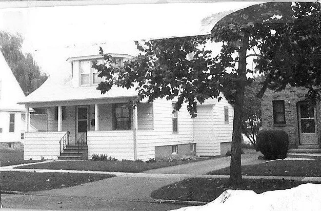 Black-and-white photo of a two-story white house with a covered front porch, sidewalk and a tree in the front yard on a residential street.