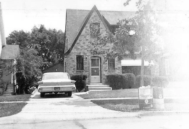 Black-and-white photo of a brick two-story house with a steep gabled roof, a vintage car parked in the driveway, and two trash barrels at the curb.