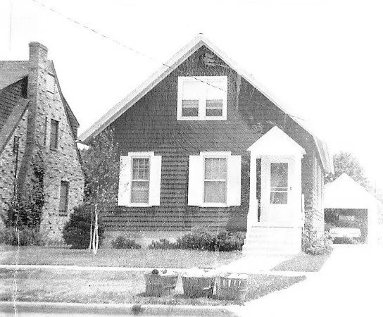 Single-family gable-front house with dark siding and white trim, small covered entry with steps, detached garage at right, stone house at left, two trash bins on the lawn.