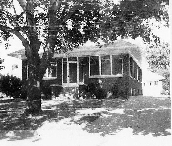 Black-and-white photo of a single-story brick bungalow with an enclosed front porch, large tree in the front yard, and a driveway leading to a detached garage.