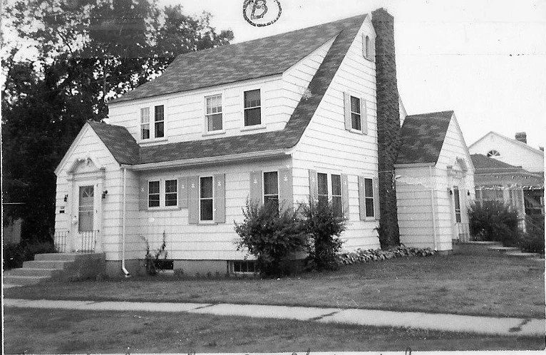 Black-and-white photo of a two-story gable-roof suburban house with clapboard siding, front steps, chimney, and a lawn.