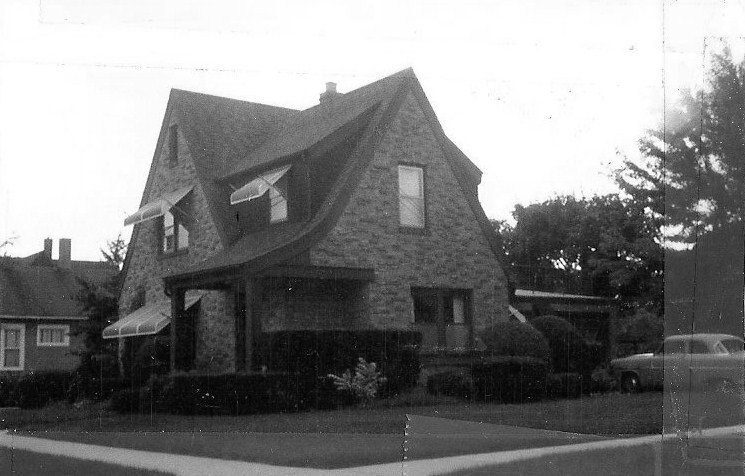 Black-and-white photo of a two-story stone house with steep gabled roof, dormer windows, front porch, shrubs, and a car parked to the right.