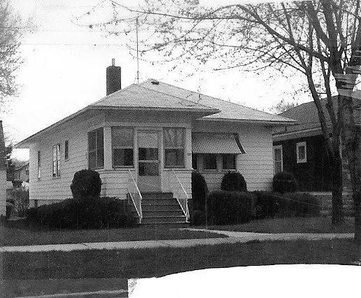 Black-and-white photo of a single-story bungalow with front steps, a small enclosed porch, an awning over a front window, and trimmed shrubs along the walkway.