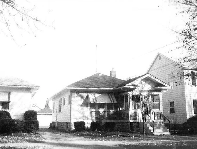 Black-and-white photo of a single-story bungalow with a small front porch, awning, front steps and shrubs, flanked by neighboring houses.