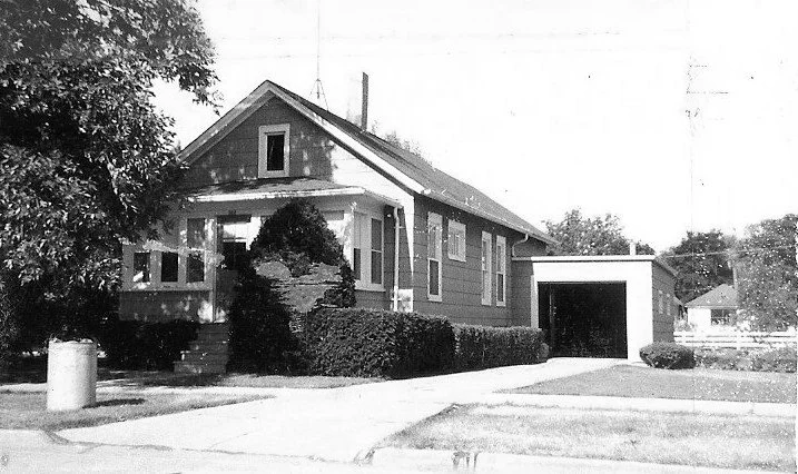 Black-and-white photo of a single-story bungalow with a front porch, shrubs and trees, a driveway leading to a detached garage on the right.