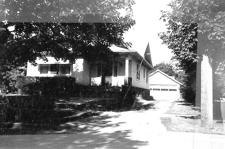 Black-and-white photo of a single-story house with a front porch and flag, driveway leading to a detached two-car garage, shaded by large trees.