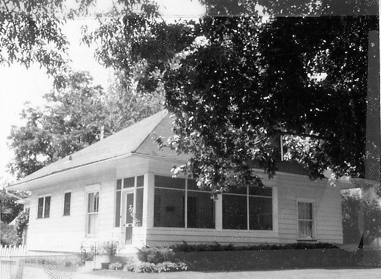 Black-and-white photo of a single-story wood-frame house with a screened front porch, large overhanging tree, small lawn and a short picket fence.