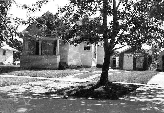 Black-and-white photo of a small bungalow with a front porch, large shade tree in the front yard, sidewalk, and a detached garage.