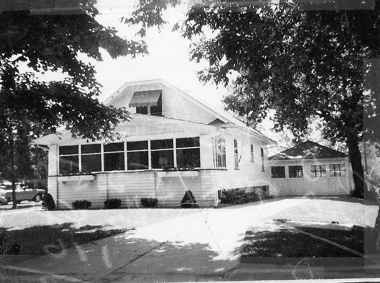 Black-and-white photo of a single-story house with an enclosed front porch, driveway leading to a detached garage, and large shade trees.