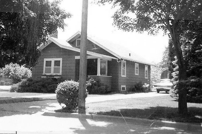 Black-and-white photo of a single-story bungalow with a front porch, trees, a utility pole, and a parked car.