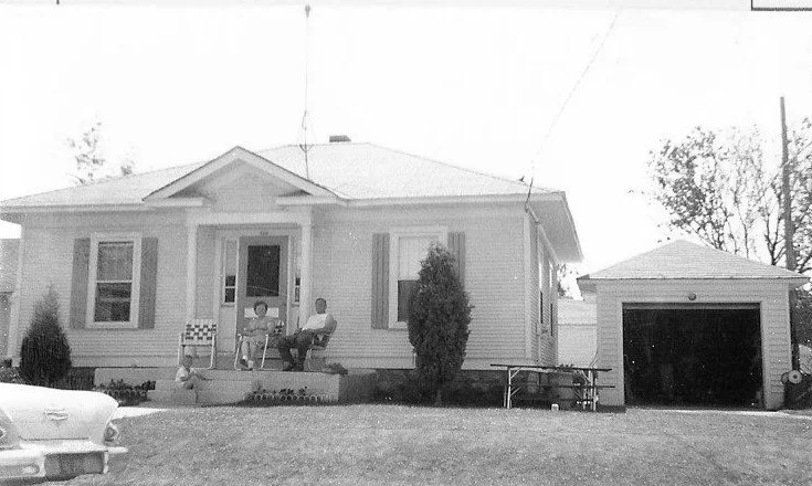 Black-and-white photo of a single-story bungalow with a small front porch where two people sit in lawn chairs, a detached garage to the right, and the front of a vintage car visible at left.