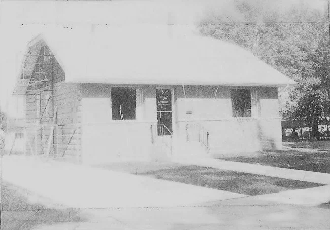 Faded black-and-white photo of a small single-story building with a central entrance flanked by two windows, scaffolding at the left and trees at the right.