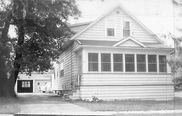 Black-and-white photo of a two-story wooden house with an enclosed front porch, driveway at left leading to a detached garage and a large tree beside the driveway.