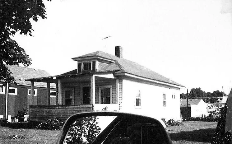 Black-and-white photo of a small single-story house with a covered front porch and a car's side-view mirror visible in the foreground.