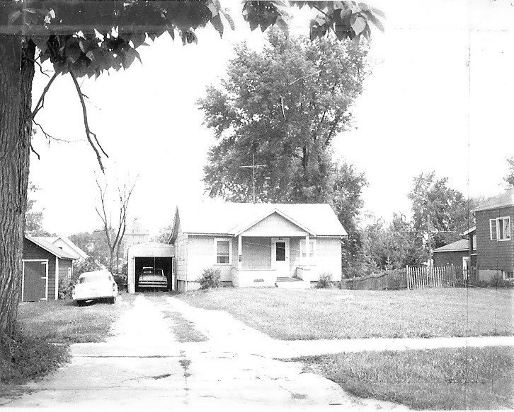 Black-and-white photo of a small single-story house with a front porch, driveway and carport with two cars, and a large tree behind.
