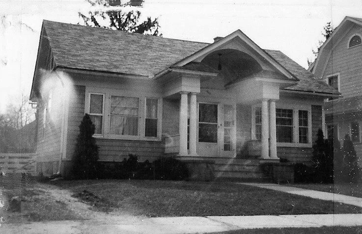 Black-and-white photo of a one-story bungalow with a covered front porch supported by two columns and a small lawn in front.