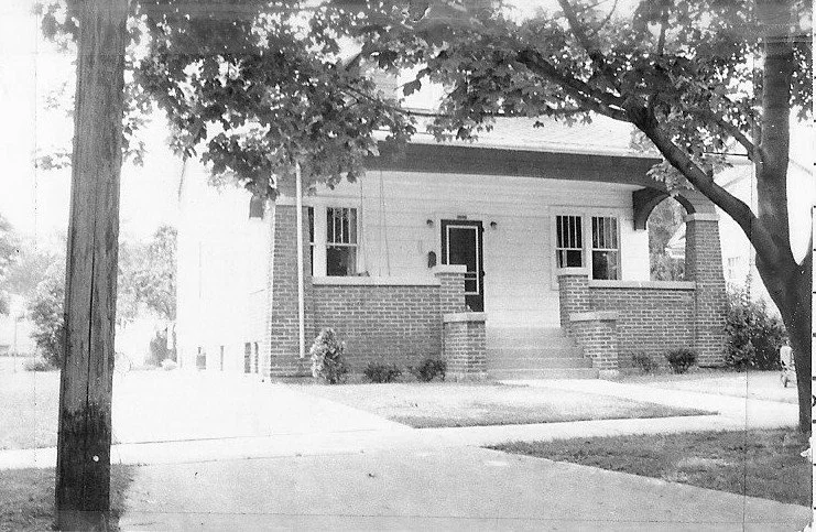 Black-and-white photo of a single-story brick-and-wood bungalow with a small front porch and steps, framed by trees and a sidewalk/driveway in the foreground.