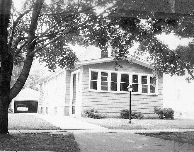Black-and-white photo of a single-story bungalow with a front enclosed porch, driveway leading to a detached garage, a lamppost in the yard, and large trees framing the scene.