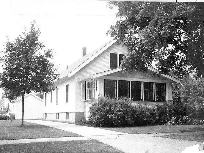 Black-and-white photo of a single-story bungalow with an enclosed front porch, driveway, detached garage, and trees in the yard.