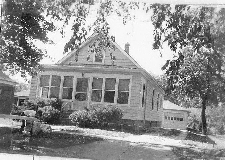 Black-and-white photo of a small bungalow with an enclosed front porch and detached garage, trees framing the scene and a person bending near the left foreground.