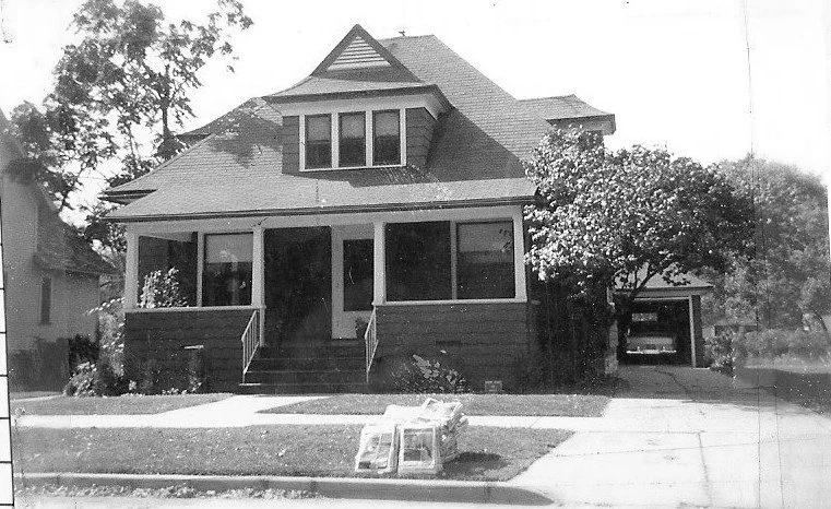 Black-and-white photo of a two-story Craftsman-style house with an enclosed front porch, tree by the driveway, a car in a detached garage, and stacked packages near the curb.