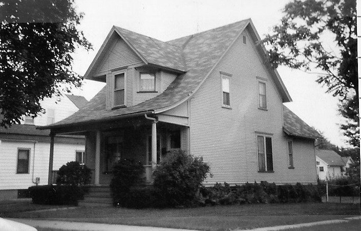 Black-and-white photo of a two-story wood-frame house with a steep gable roof, front porch, and shrubs along the yard.