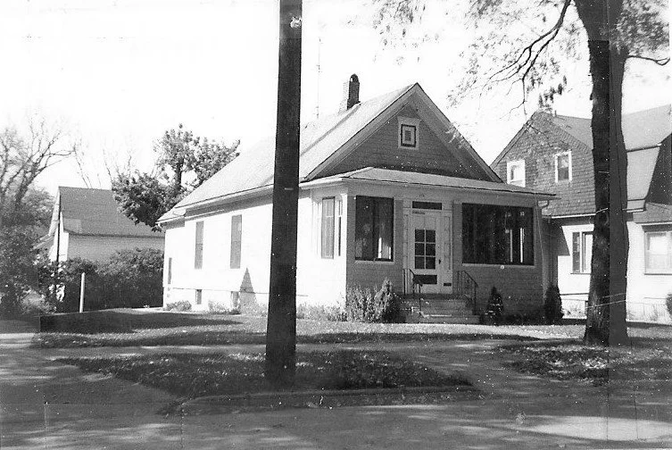 Black-and-white photo of a single-story bungalow with a small front porch, trees in the yard, and neighboring houses.