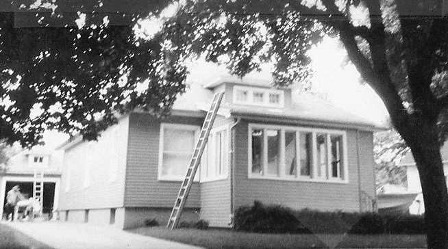 Black-and-white photo of a single-story house with a ladder leaning to the roof, a tree in the foreground and a person near an open garage in the background.
