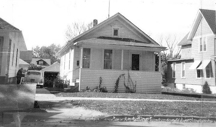 Black-and-white photo of a small single-story bungalow with front porch, driveway with an old car, and a person beside the car.